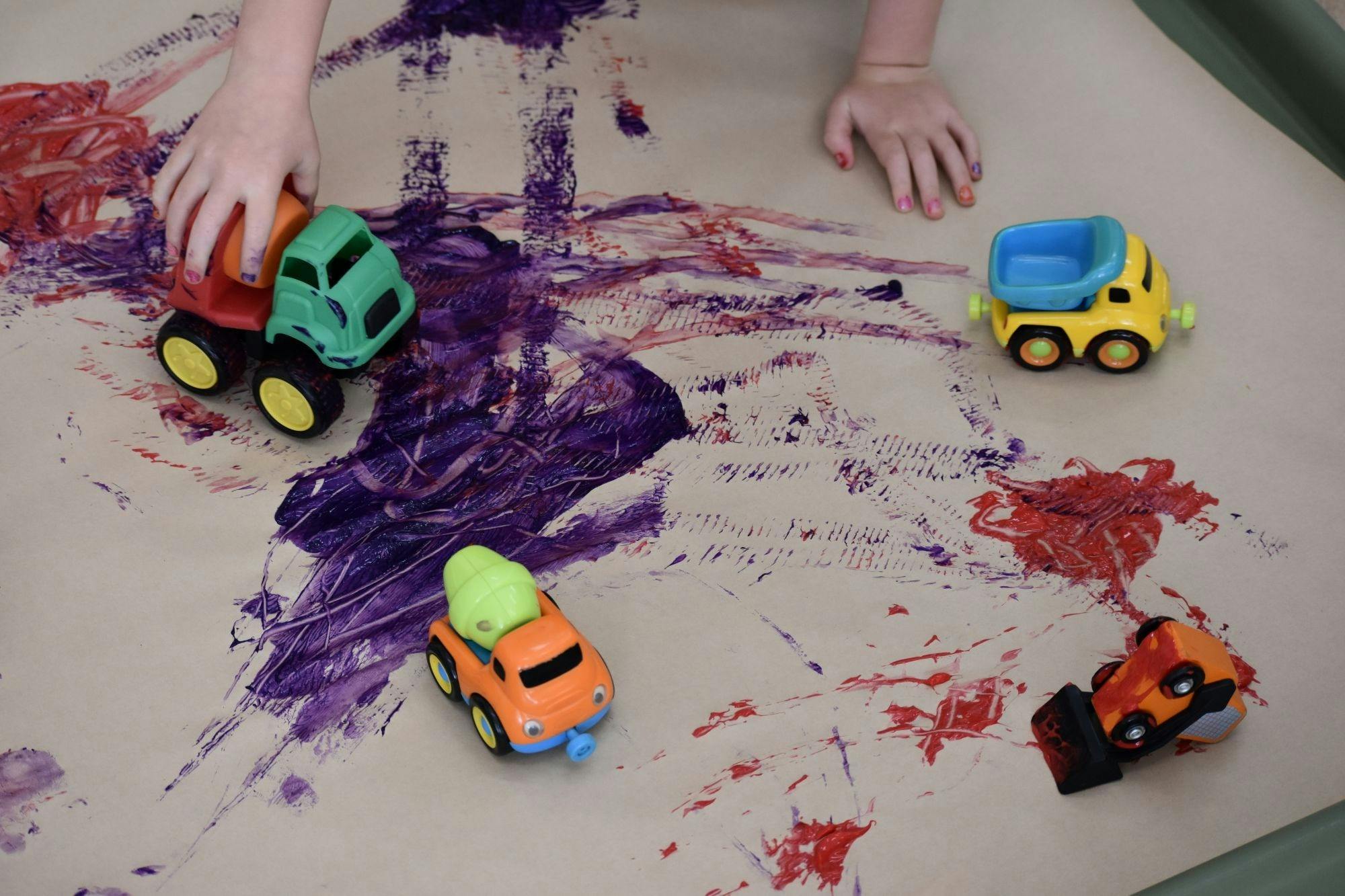 Children's hands pushing toy cars through paint on a sheet of paper to make track prints