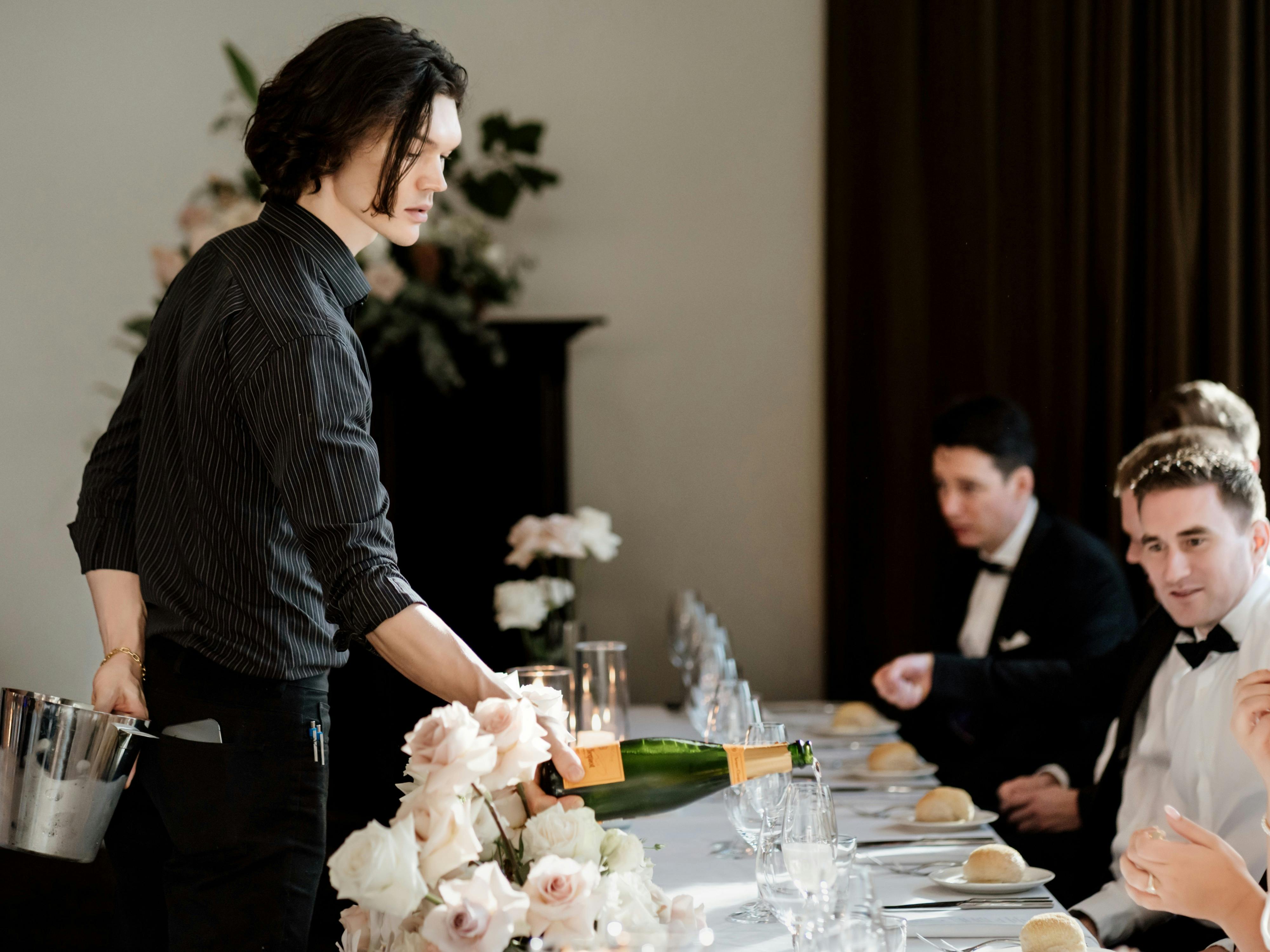 Guests at a wedding table with beautiful flowers and table setting served champagne by waiter.