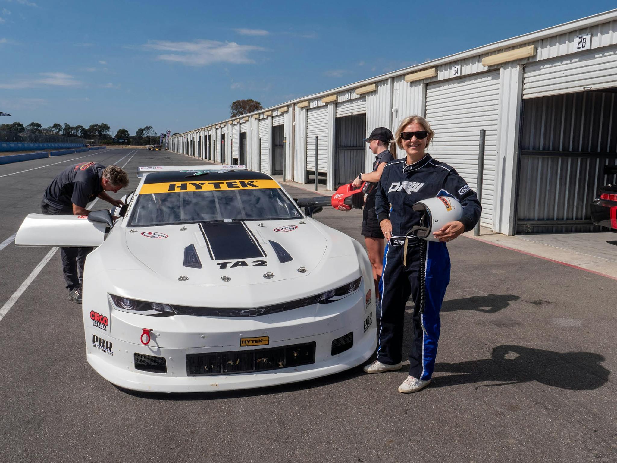 Lady in a racing suit standing next to a TA2 white muscle car