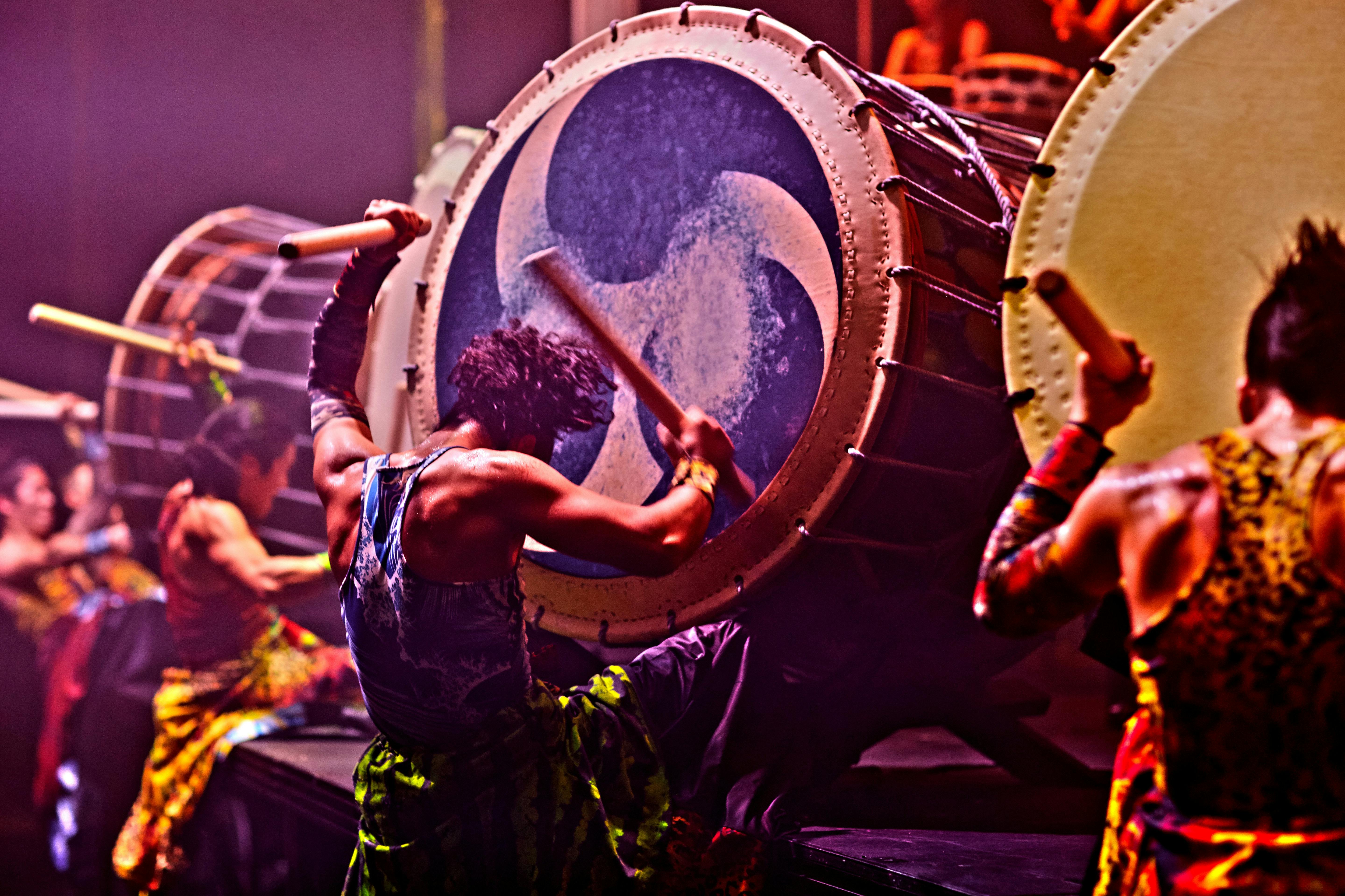 Back view of four drummers beating large taiko drums with wooden baton style sticks.
