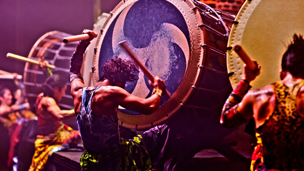 Back view of four drummers beating large taiko drums with wooden baton style sticks.