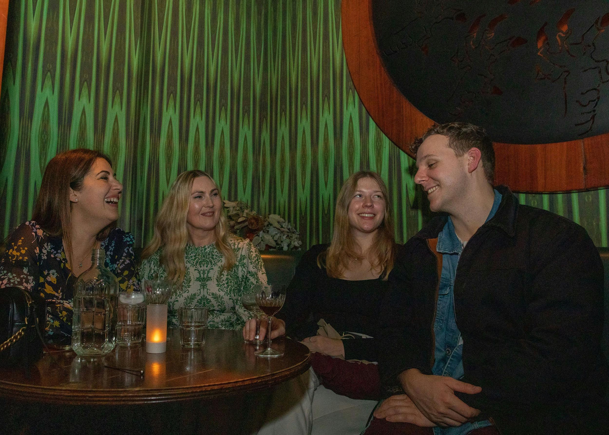 Three women and a man are sitting in a booth drinking cocktails.  The walls are green.