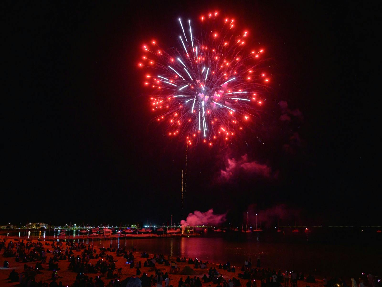 Large red fireworks bursting above a beach filled with spectators
