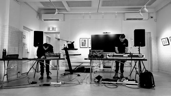 Two electronic musicians perform with synthesizers and cables in a contemporary gallery space.