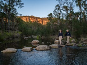 Tour guides cross the creek via stepping stones at Carnarvon Gorge, with cliffs bathed in sunlight
