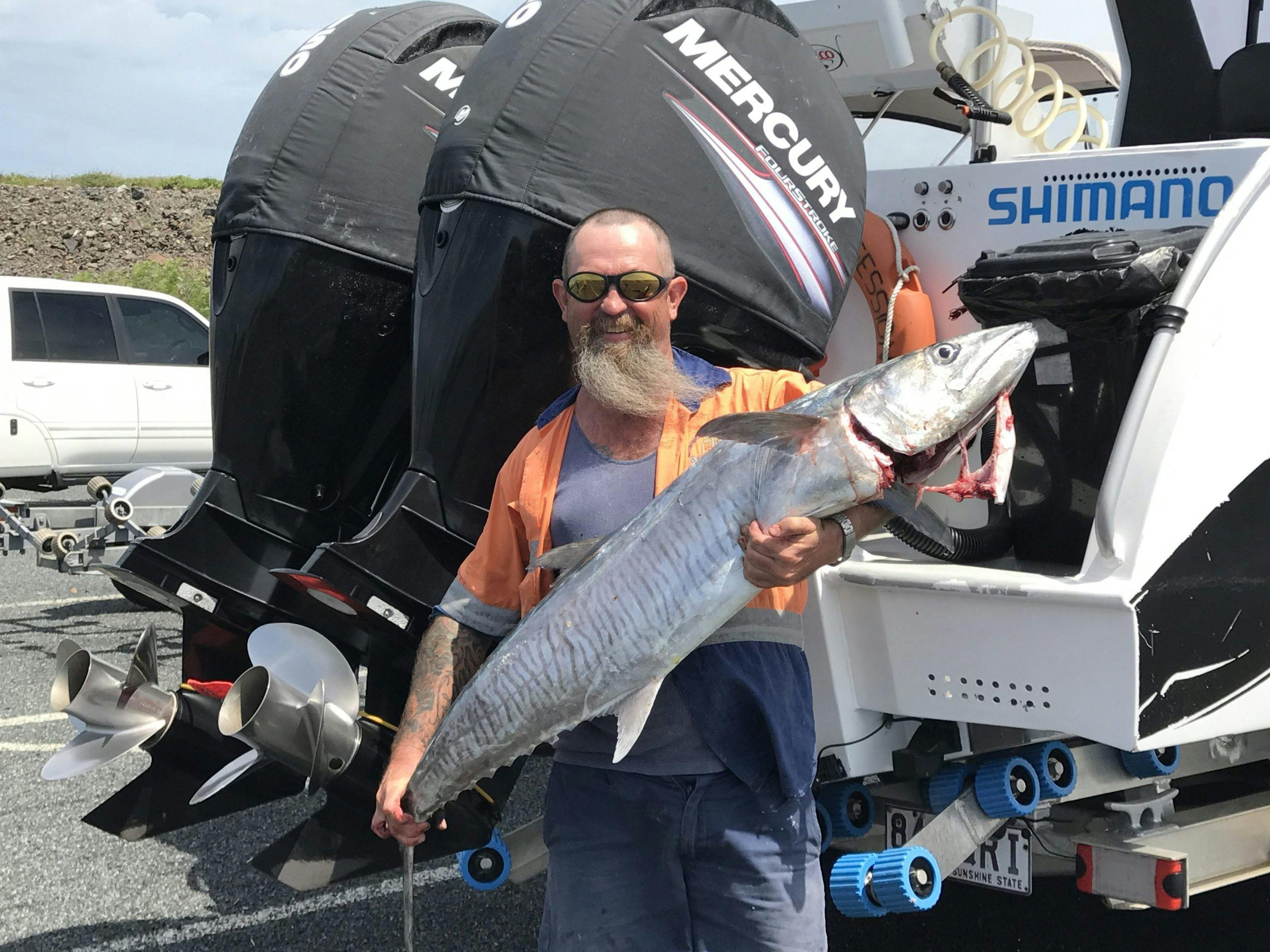Angler hold up a large Spanish Mackerel standing behind the boat. The 2 large motors are behind him.