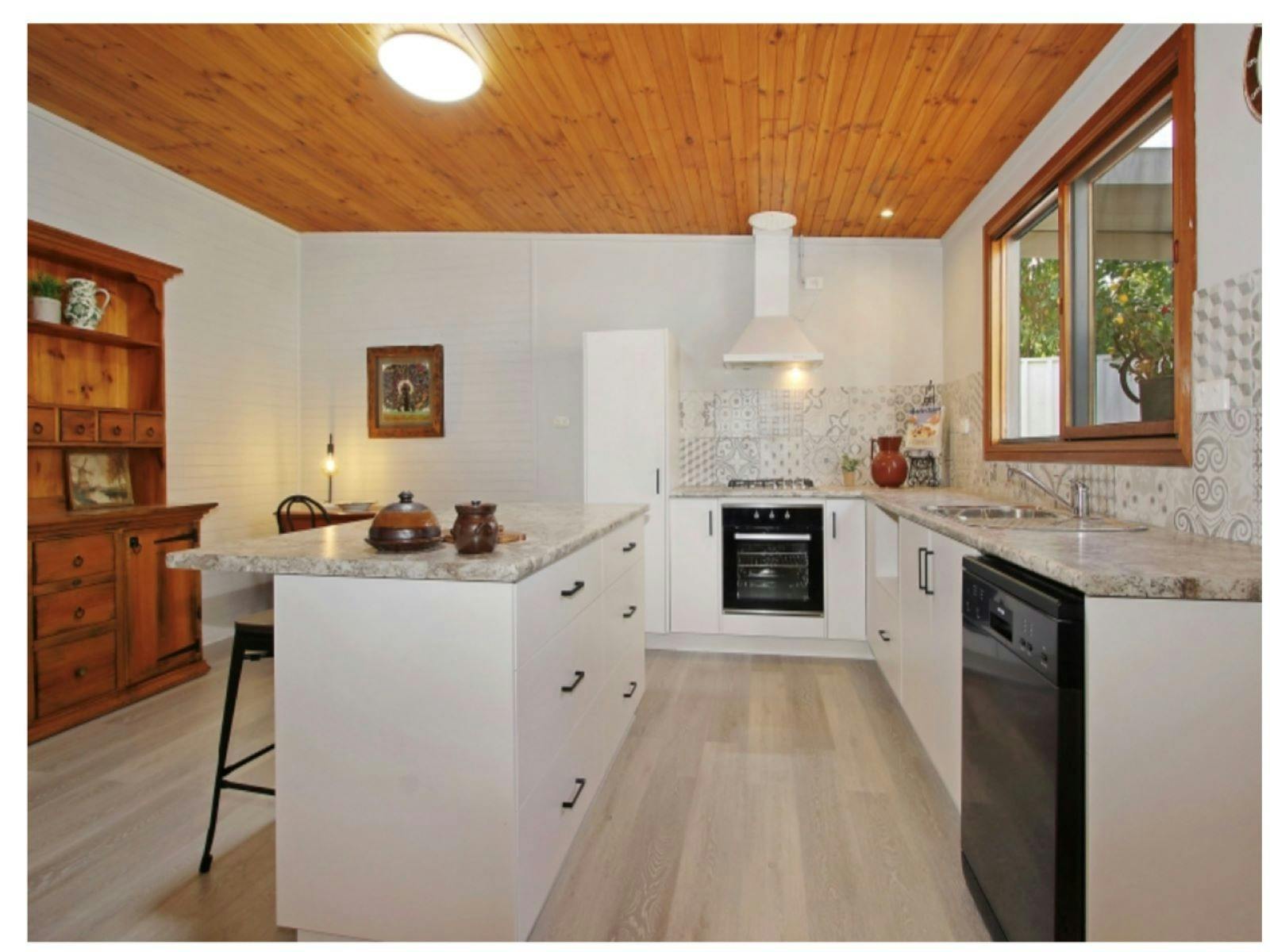 Kitchen, white cupboards with marble tops. Breakfast bench in the middle, black dishwasher and oven.