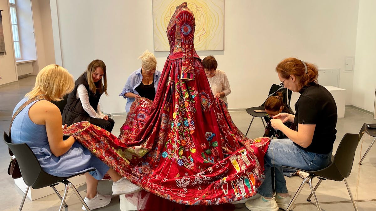 Four seated women stitching onto the skirt of a red dress