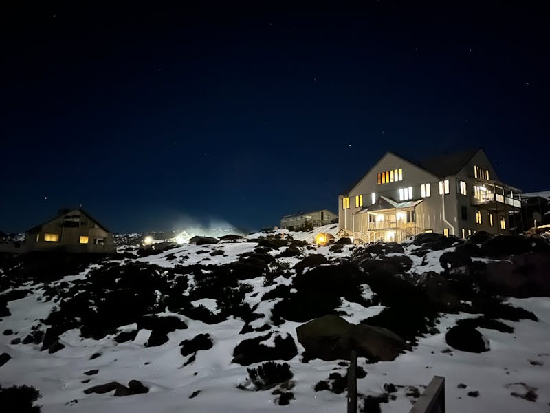 A three story ski lodge at night, with lights in the windows, a clear night sky, snow on the ground