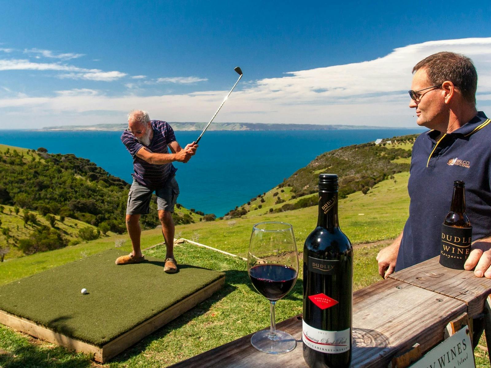 In the foreground a man enjoys a glass of wine while in the background a man swings a golf club