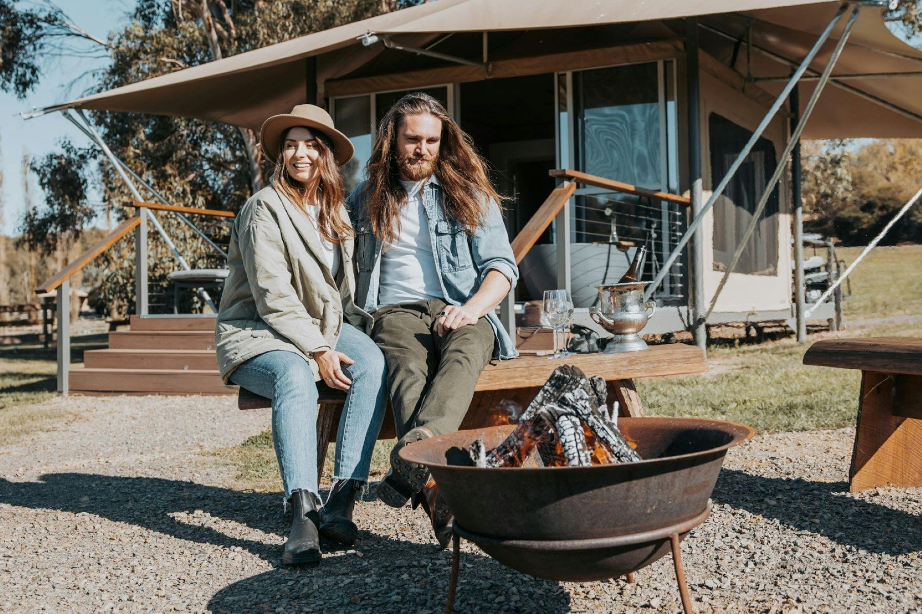 Two people sitting side by side by an open fire pit in front of an eco glamping tent.