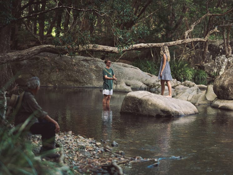 Two young people stand in the middle of a rocky creek, surrounded by trees as they fish.