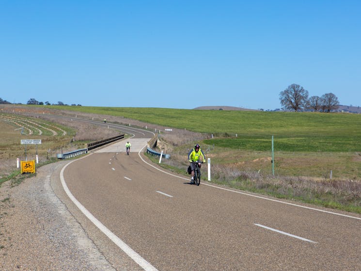 Three riders on a cycling tour pass over the Inglewood Bridge. Green fields and blue sky.