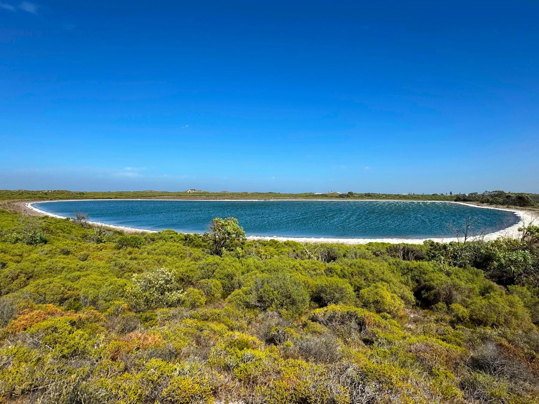 View of lake surrounded by a thin strip of white sand and green shrubs.