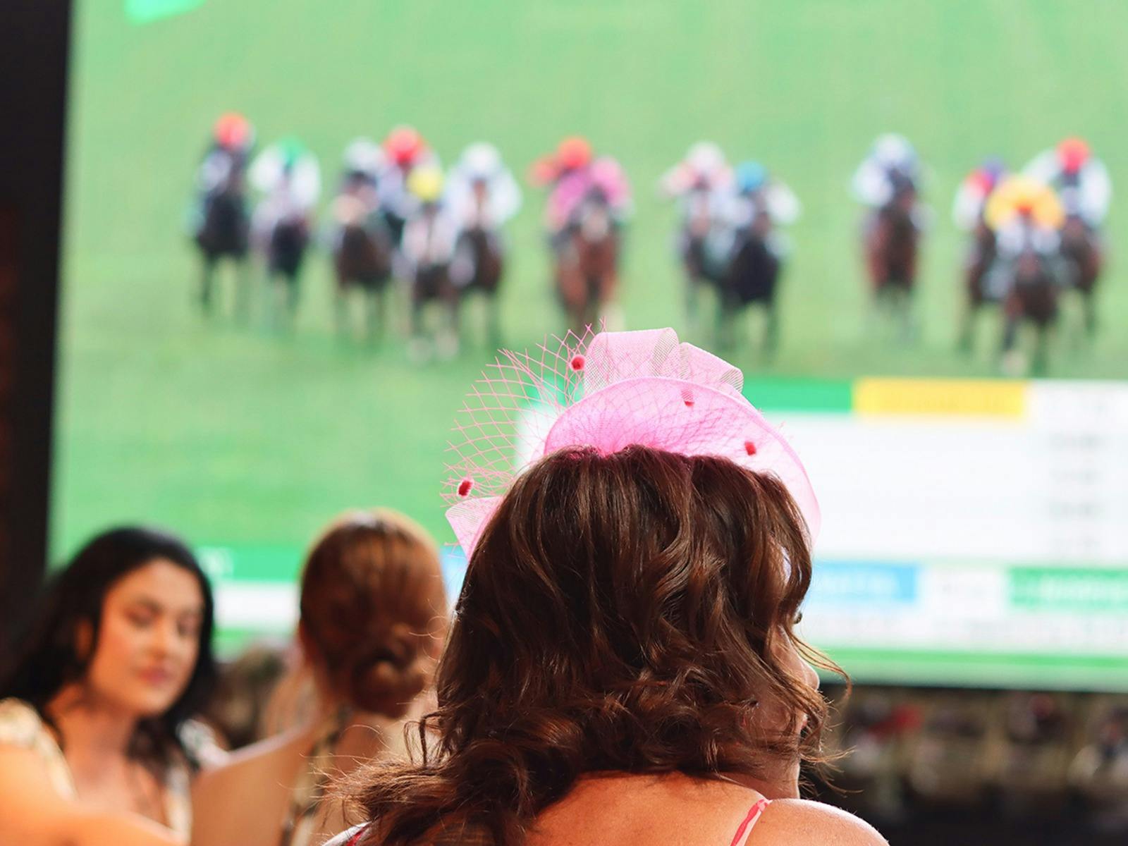 A woman in a pink fascinator watching the Melbourne Cup on a large screen