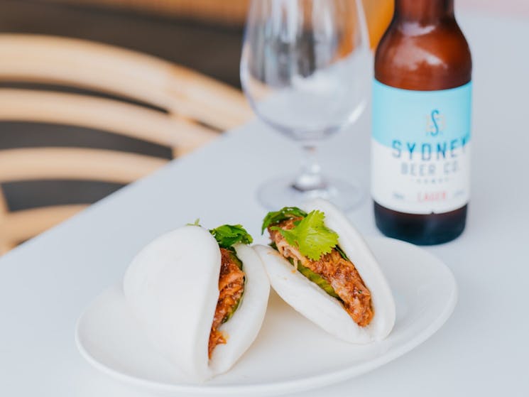Duck filled bao buns in the fore, on a white table with beer and glass in the background.