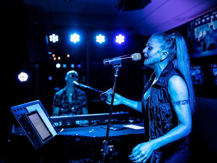 Woman in foreground singing into a mic with man in the background playing keyboard