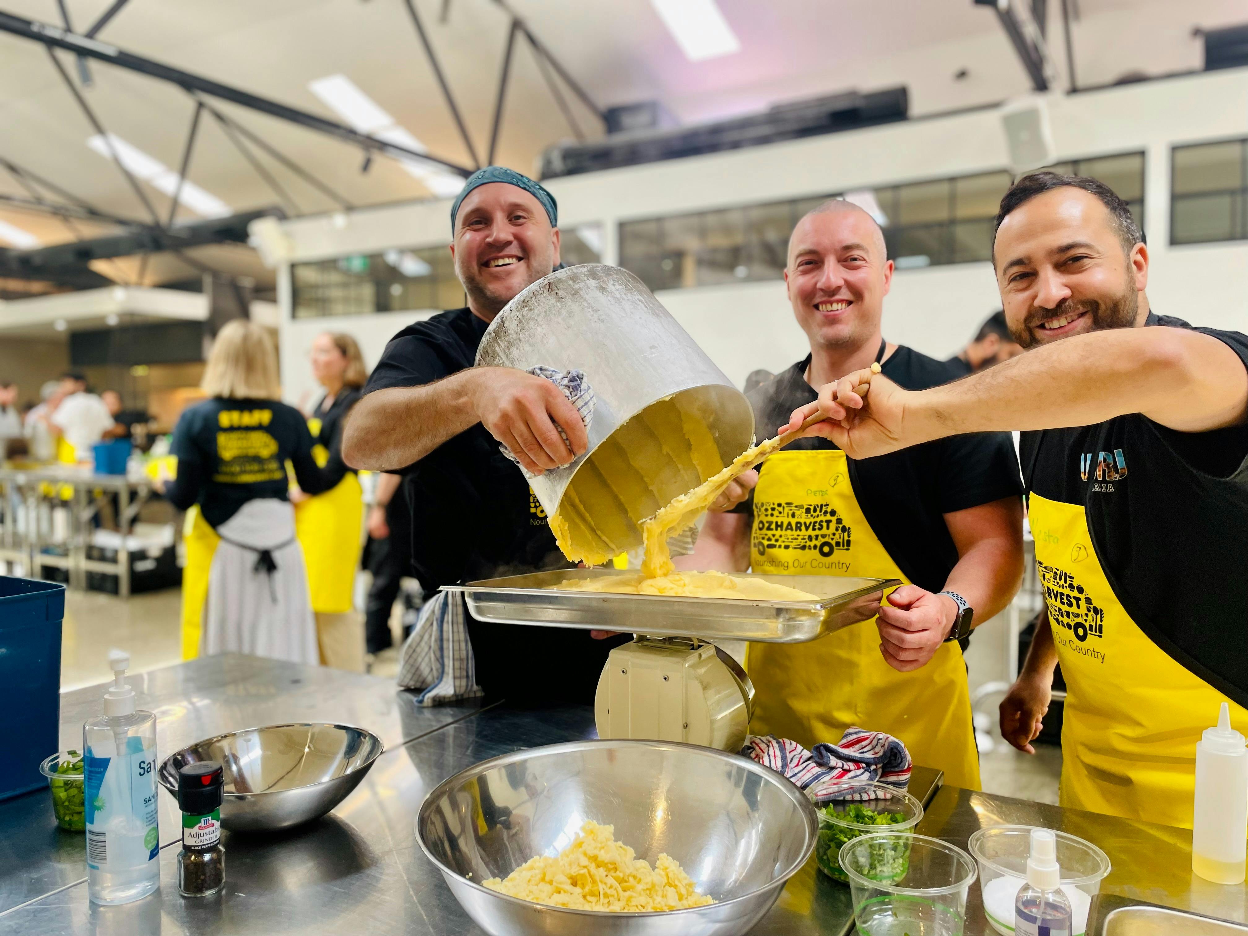 Three people laughing while pouring mashed potato from a large pot into a tray during an OzHarvest C