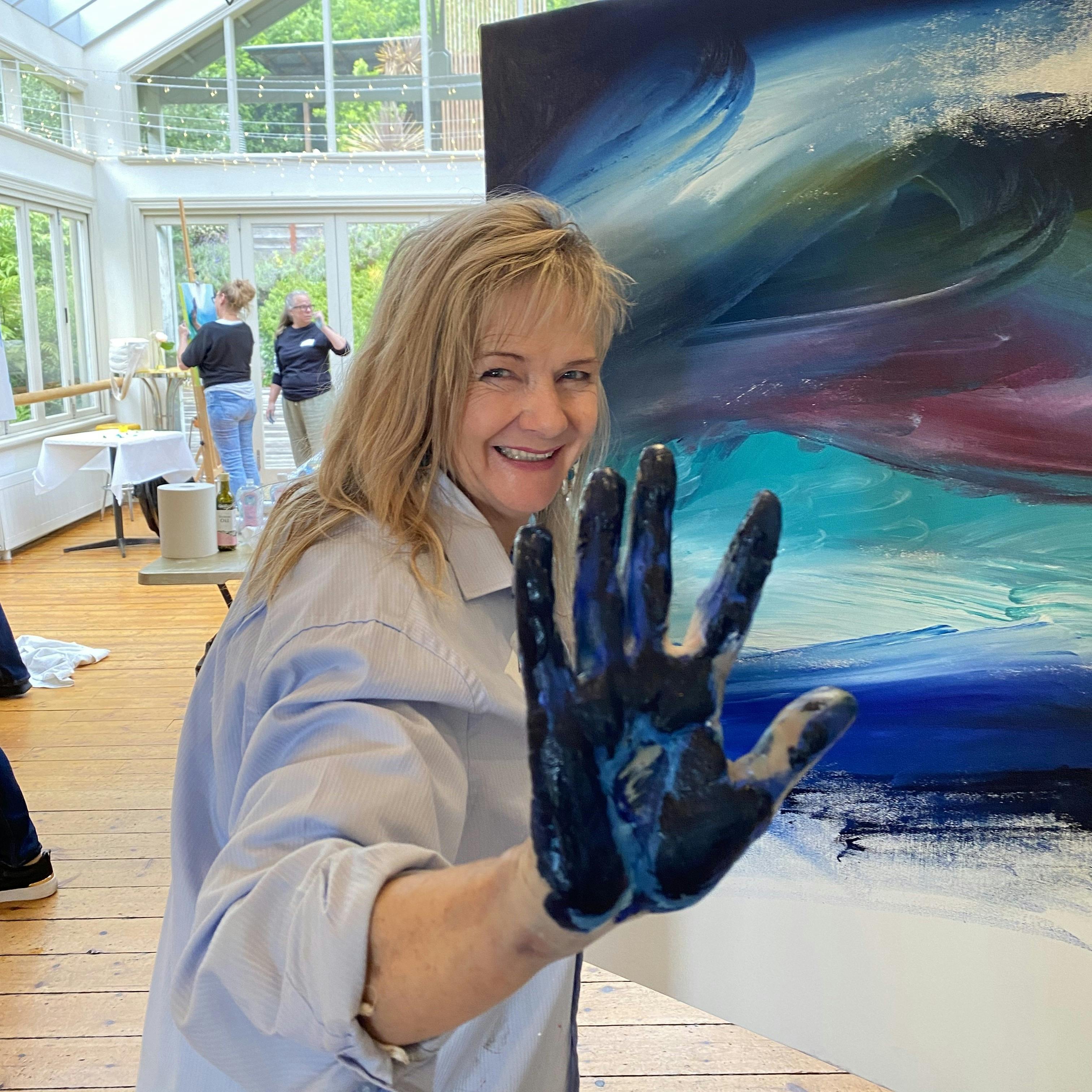 a woman smiling at the camera showing her hand that is covered in paint, standing next to her art