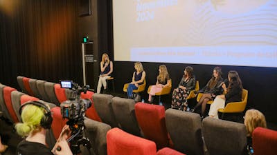 The image depicts a panel discussion taking place in front of a cinema screen