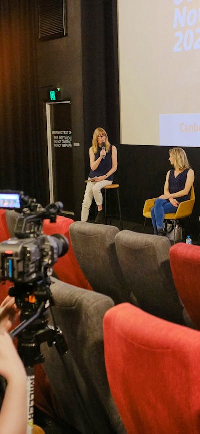 The image depicts a panel discussion taking place in front of a cinema screen
