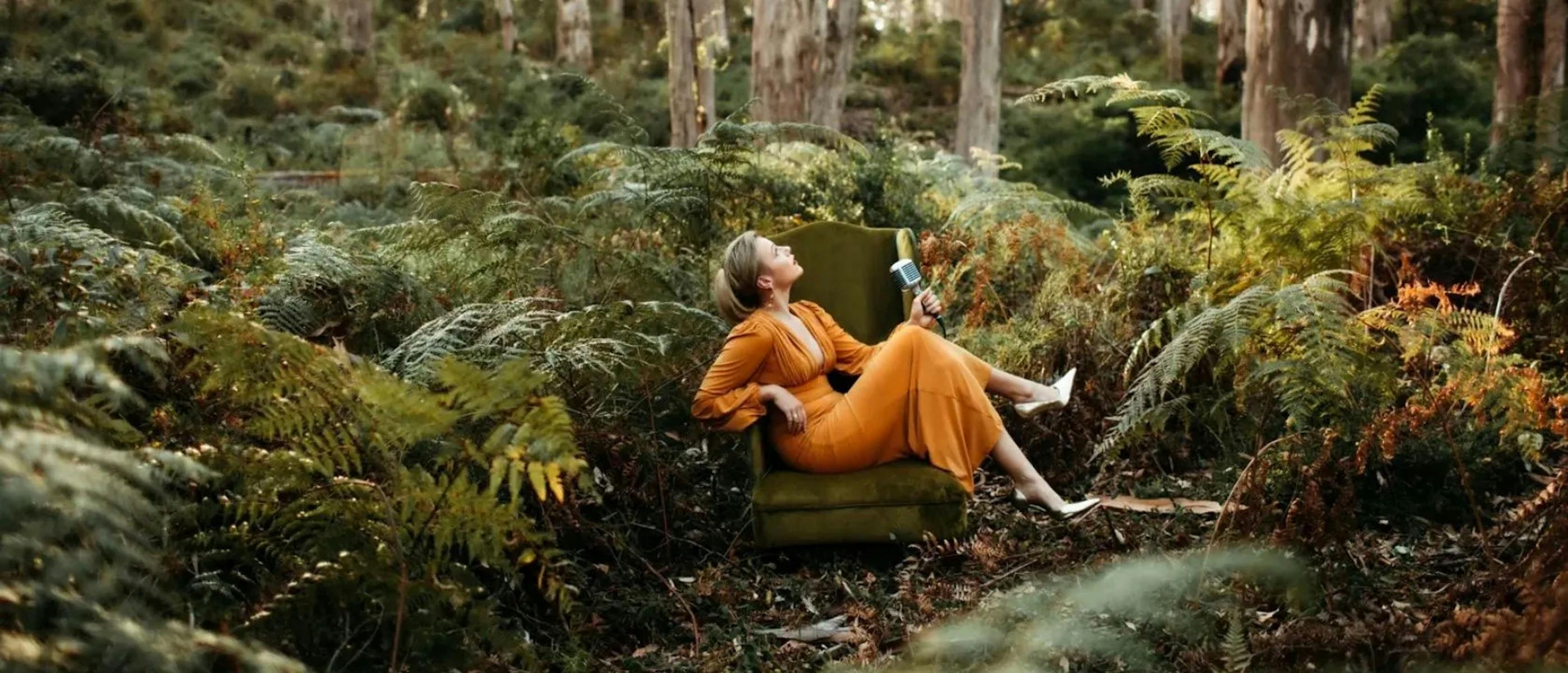 A woman wearing an orange dress holding a microphone sitting on an armchair in a forest