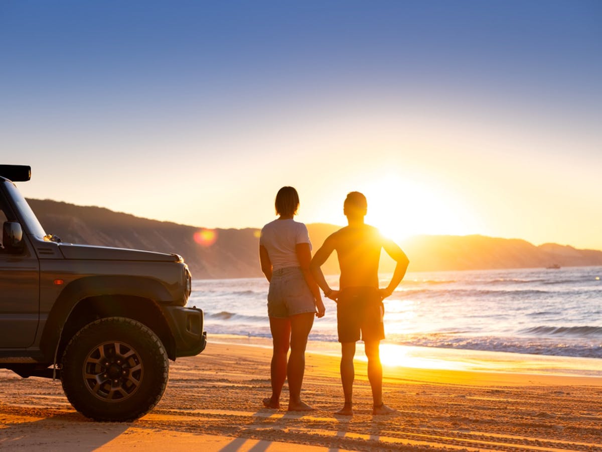A couple on the beach with their car looking out at the ocean while the sunsets