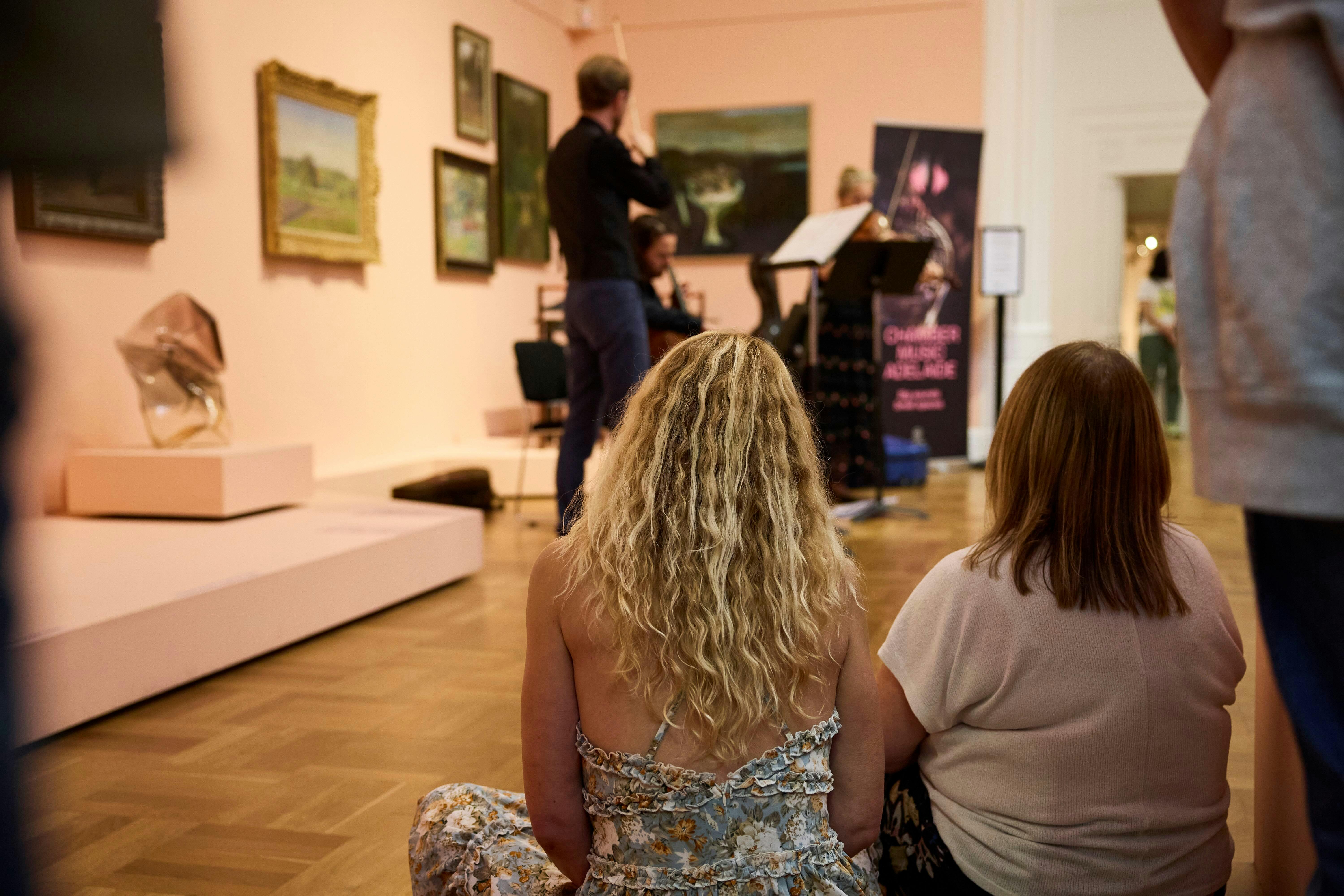 An informally gathered audience are watching a string ensemble perform in an art gallery.