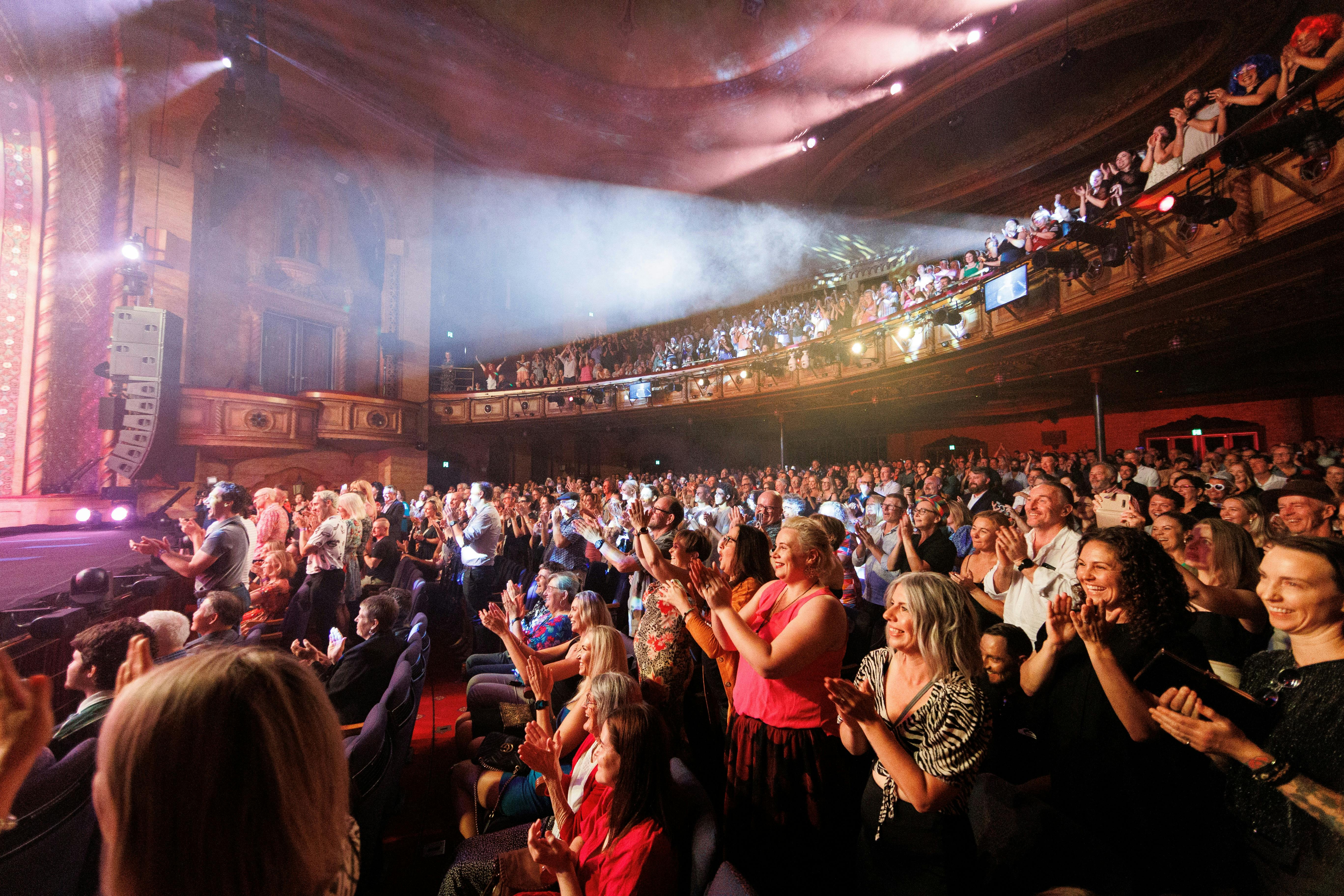 Photo of a crowd in the Civic Theatre standing to applaud. The lights are hazy with a pink hue.