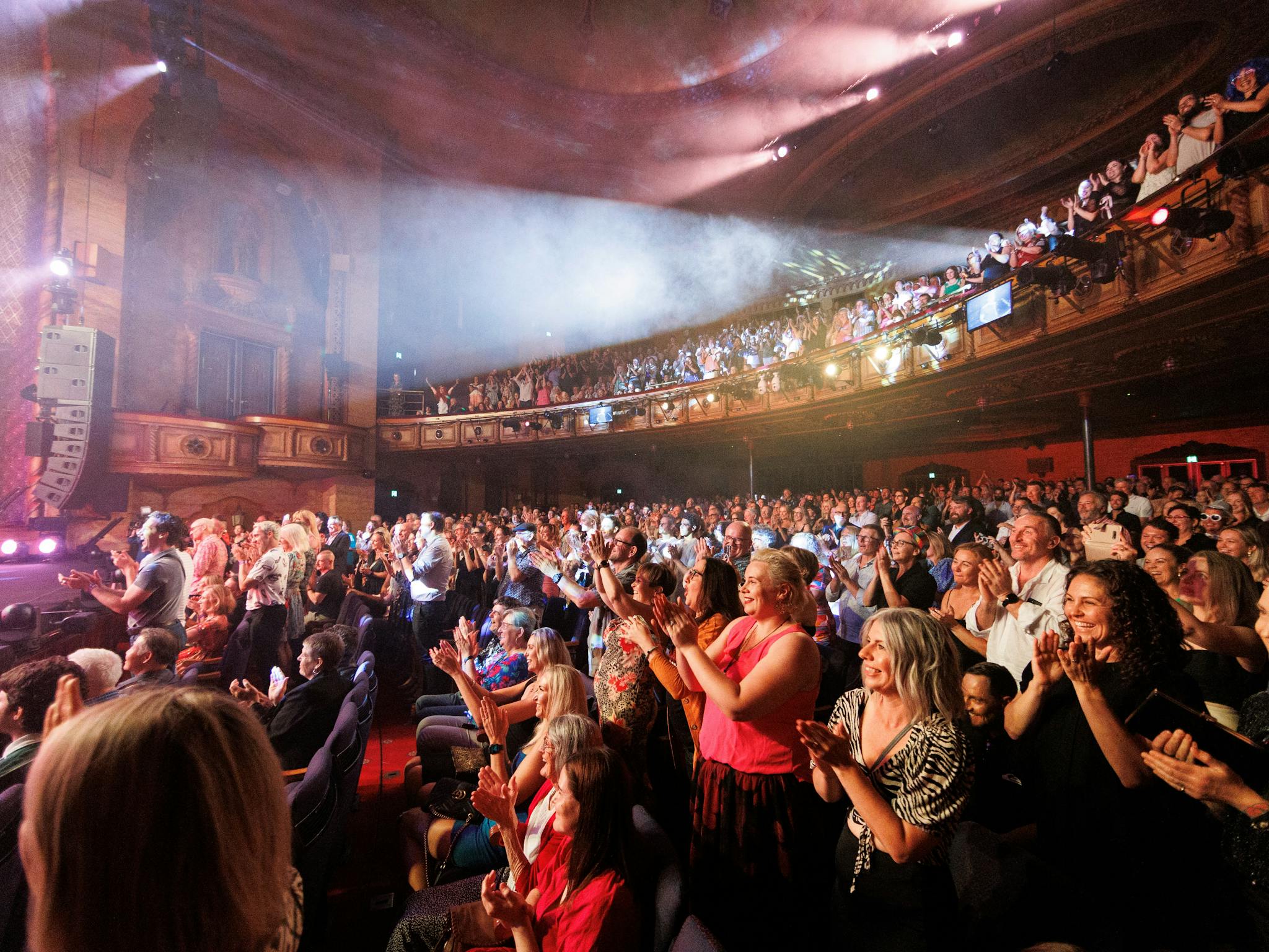 Photo of a crowd in the Civic Theatre standing to applaud. The lights are hazy with a pink hue.