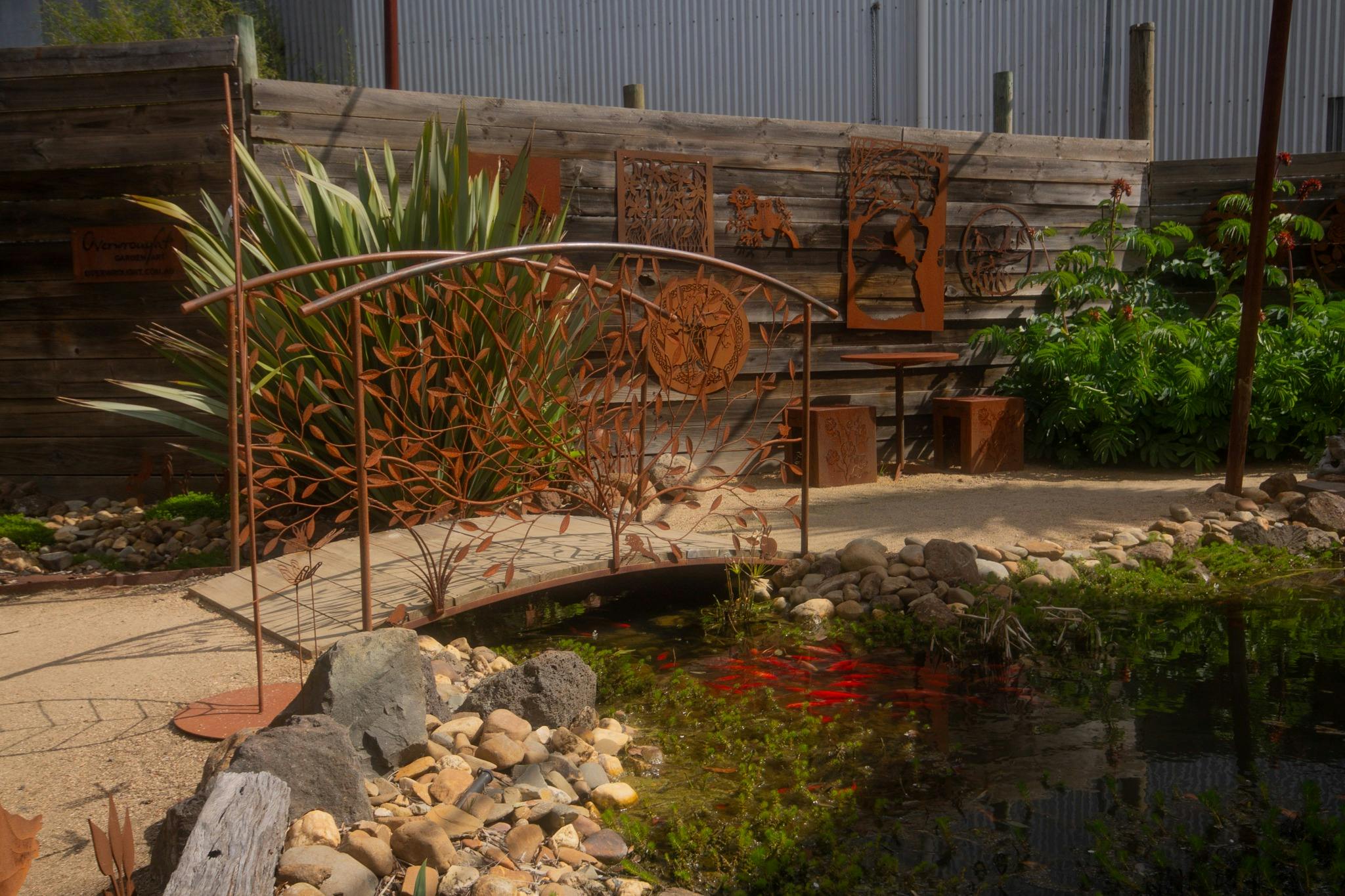 In the forground is a pond brimming with fish behind is an ornate metal bridge and range of wallart.