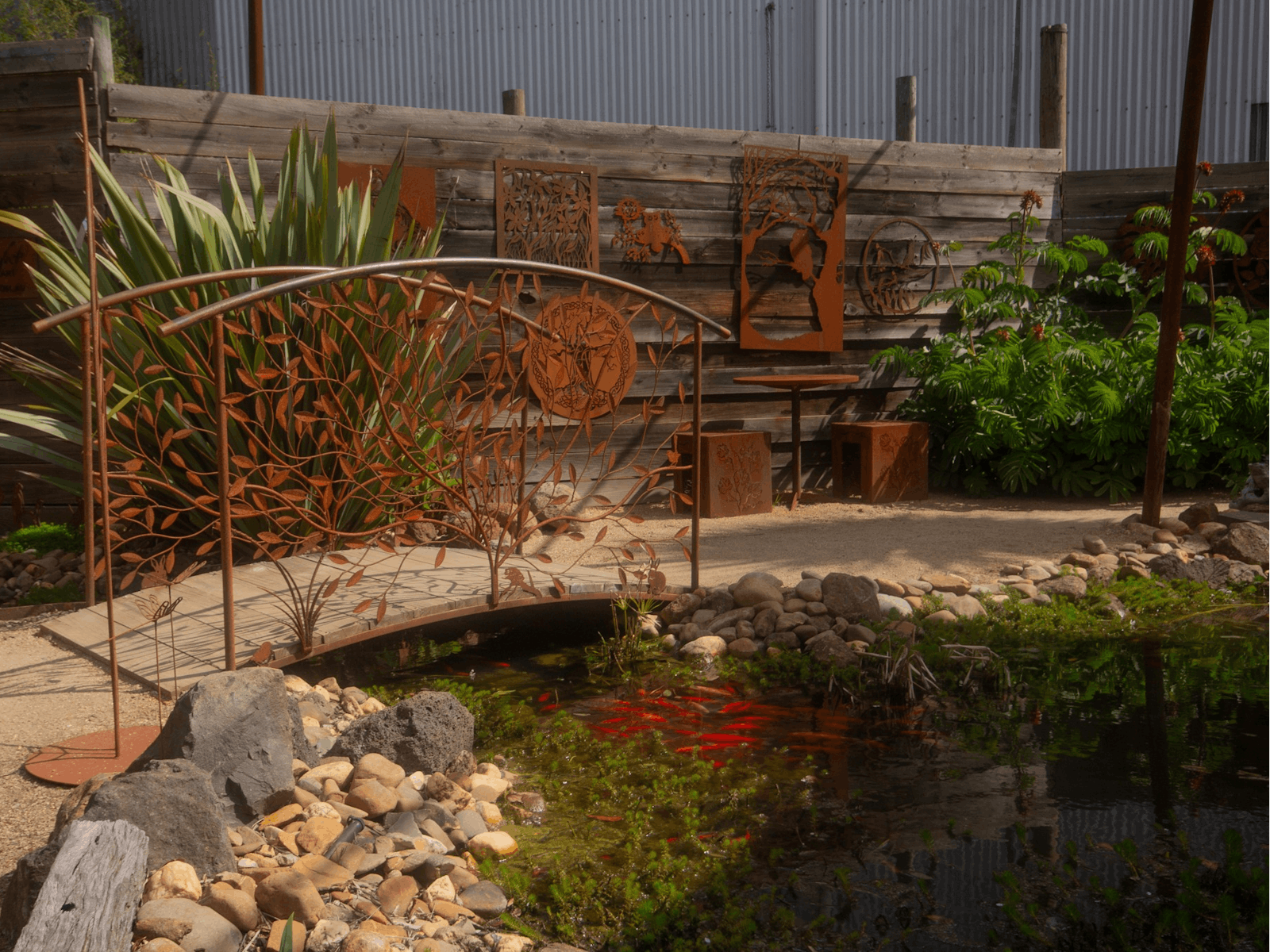 In the forground is a pond brimming with fish behind is an ornate metal bridge and range of wallart.