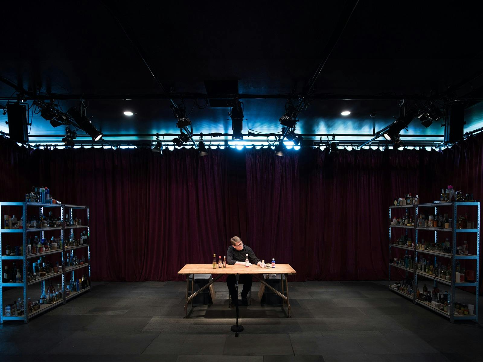 A person wearing black sits at a trestle table. They are spotlighted in a dark room.