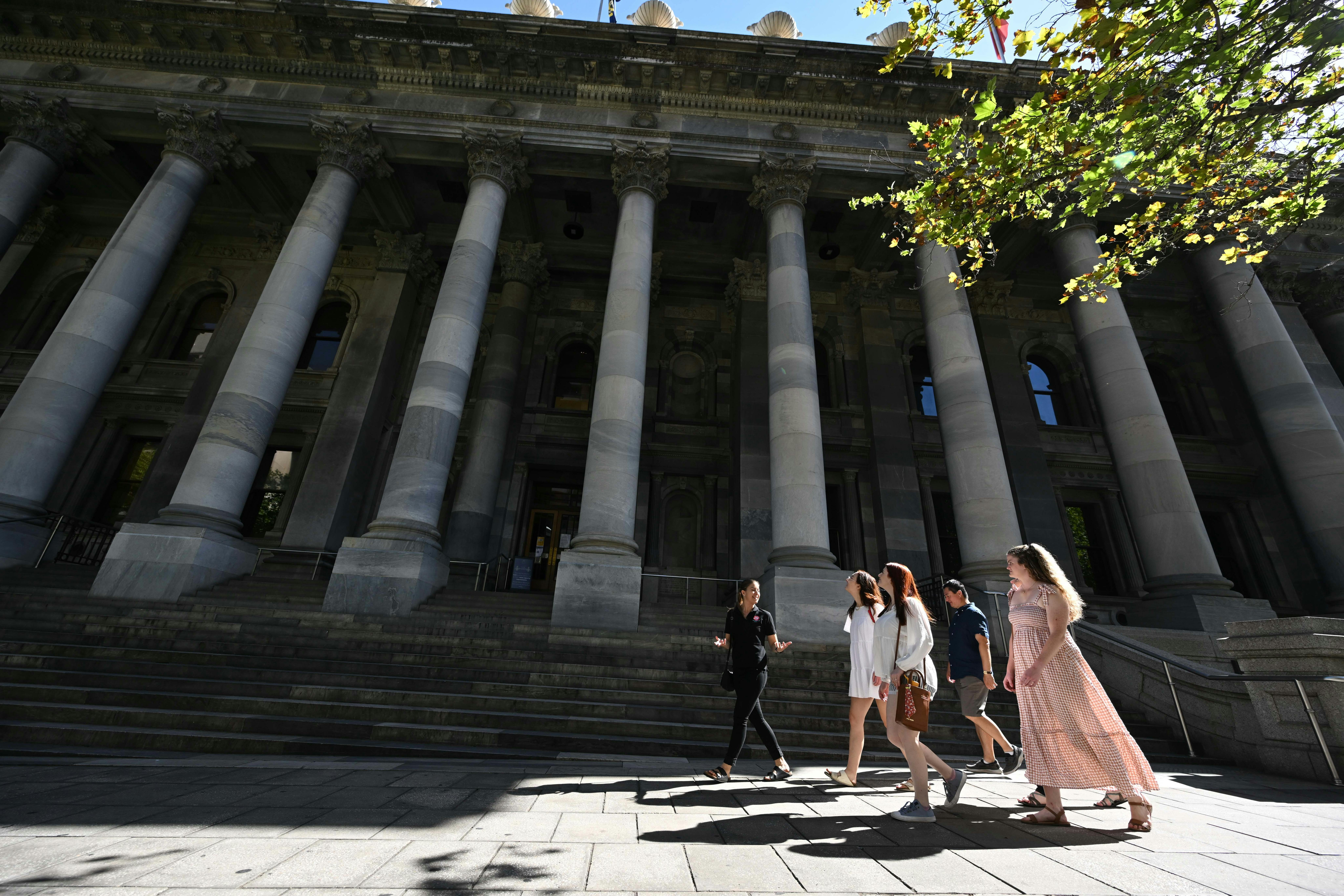A group is led by a tour guide past a grey building with marble columns.