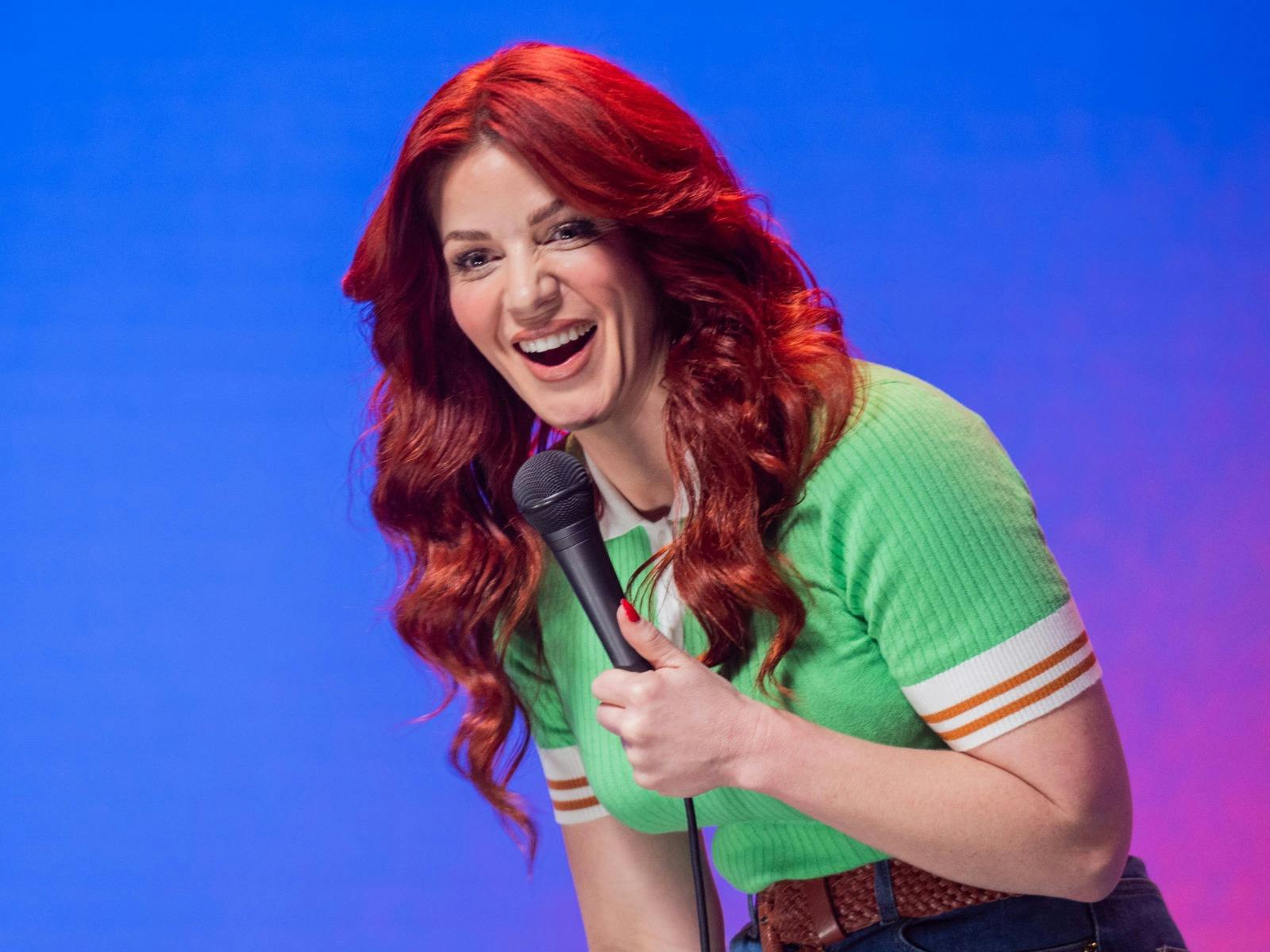 A woman with long red hair wearing a green shirt smiles and holds a microphone