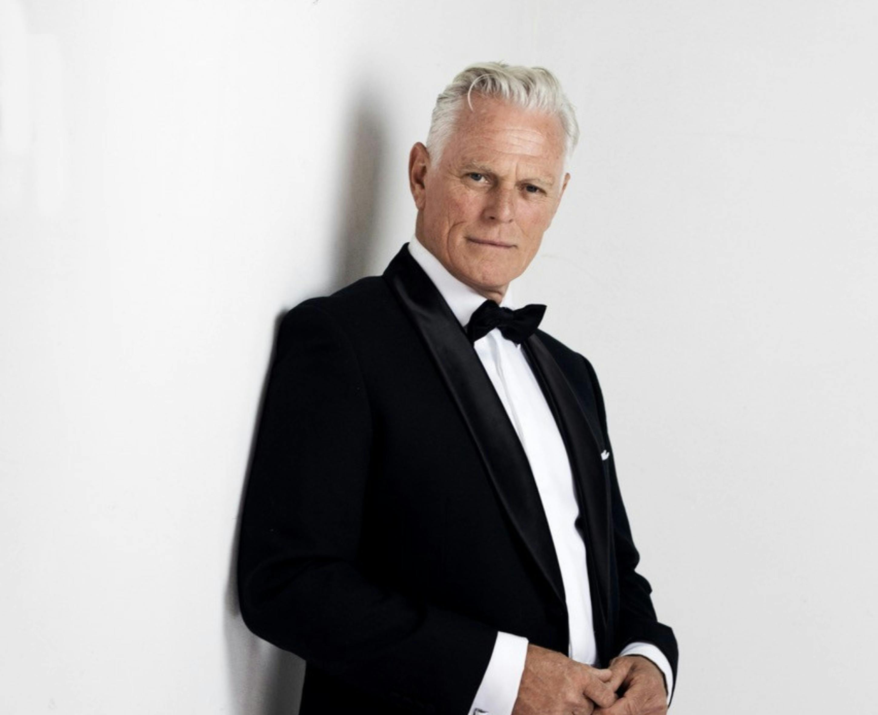 Michael Cormick in a tux leaning on a white wall smouldering