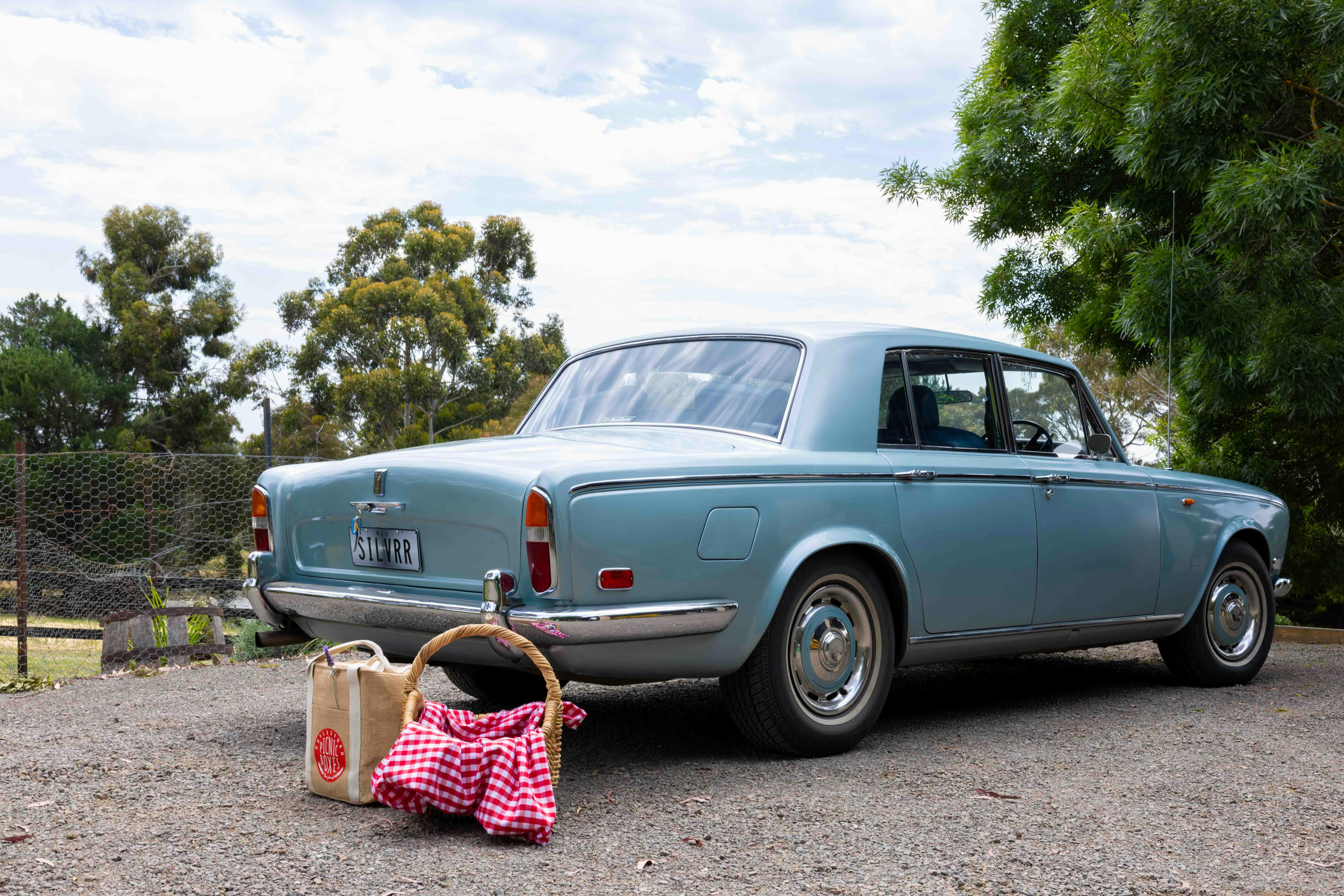 picnic baskets on the ground beside the back of a silver-blue vintage Rolls Royce
