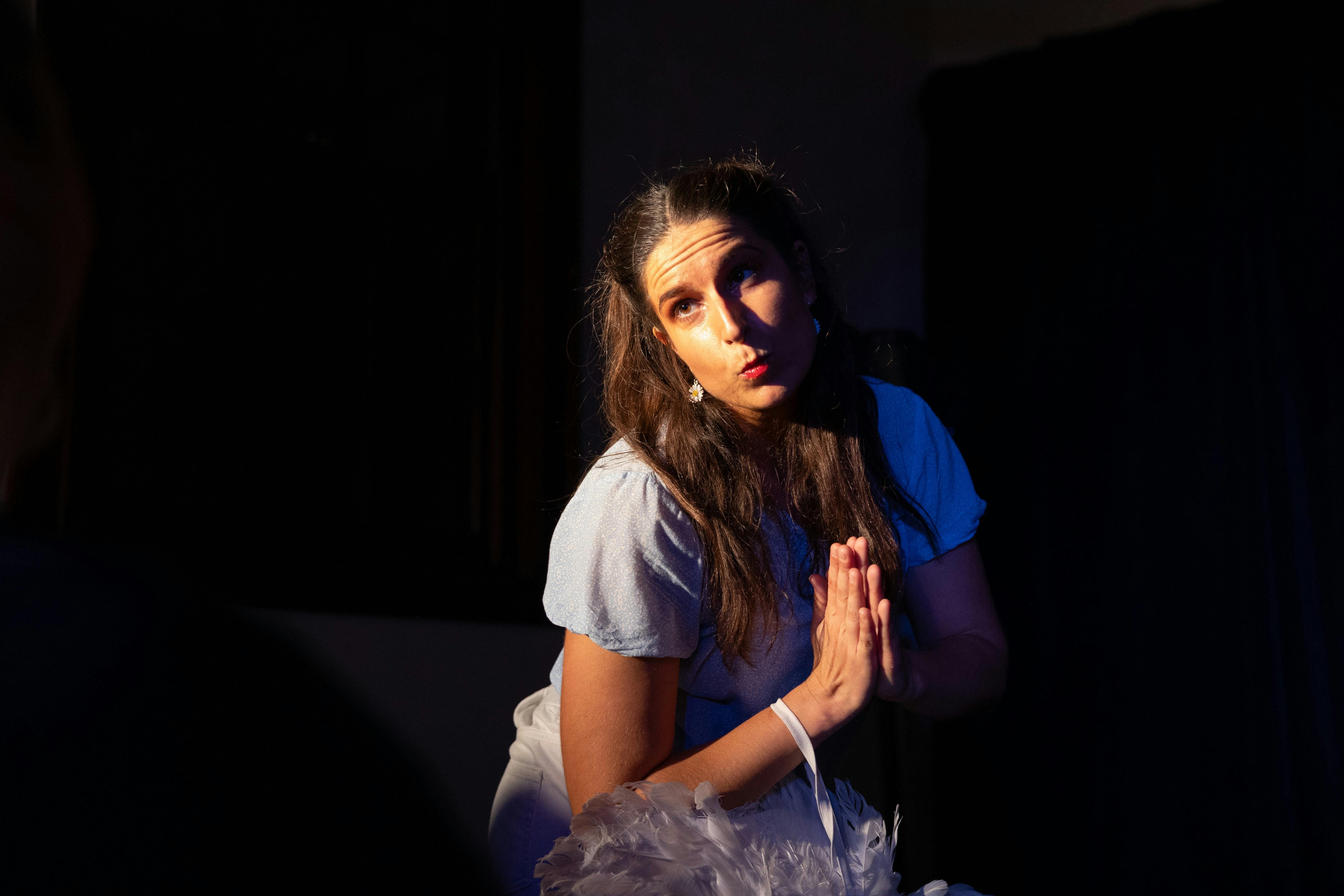 A Woman with pursed lips and prayer hands looking at the audience. Black background.
