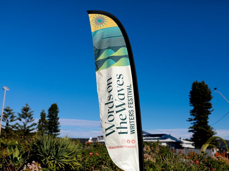 A flag with Words on the Waves Writers Festival branding is shown against a blue sky and greenery