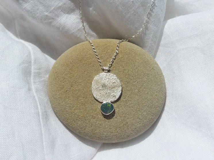A fine silver necklace with a silver circular pendant and green gemstone sitting upon a round pebble
