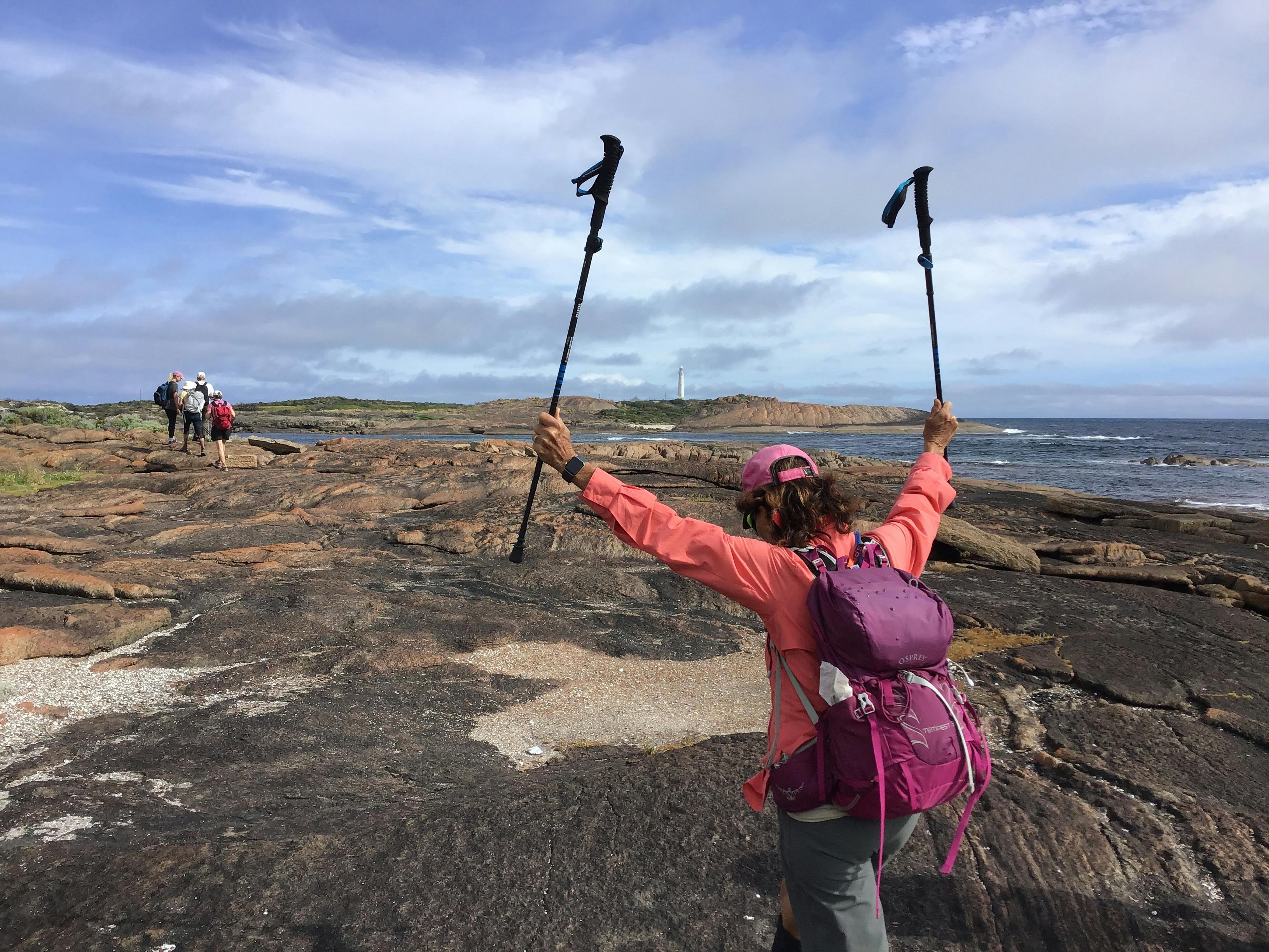 Woman hiker holds up two hiking poles in triumph, walking towards a lighthouse.