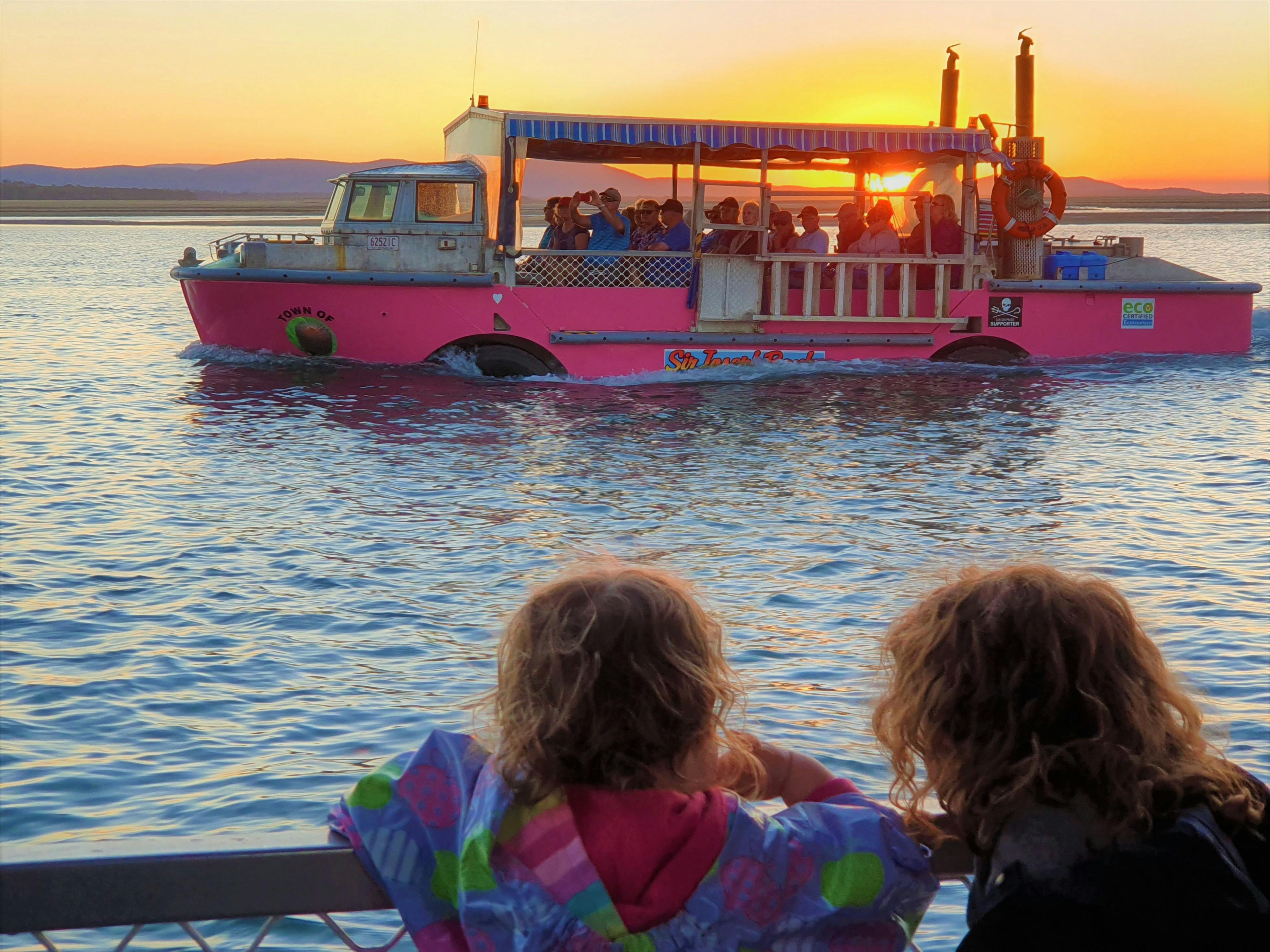 two young children watch travelling amphibious vehicle from another as the sun sets behind