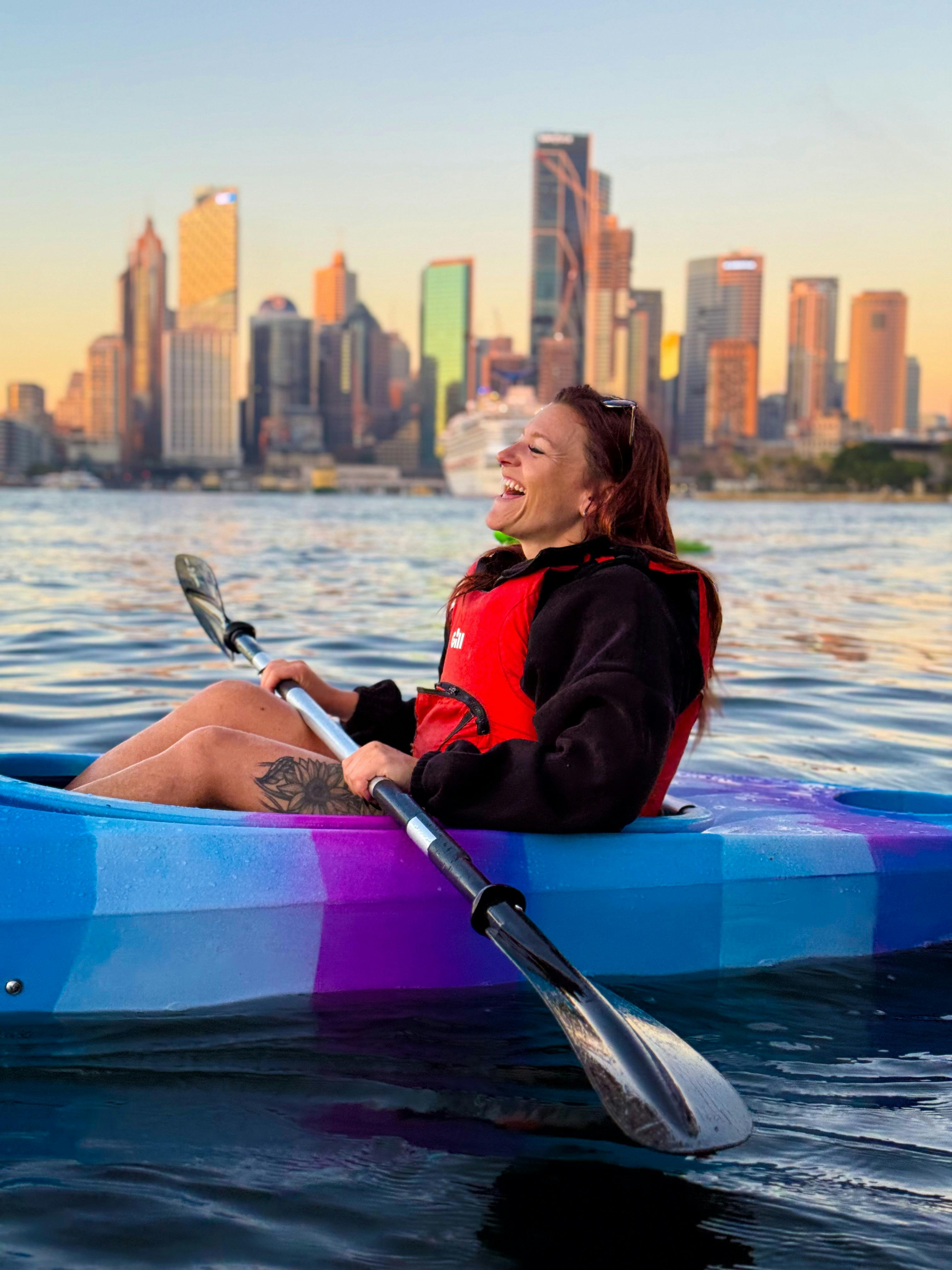 Lächelnde Dame, die in ihrem Kajak sitzt, im Hintergrund das Geschäftsviertel von Sydney