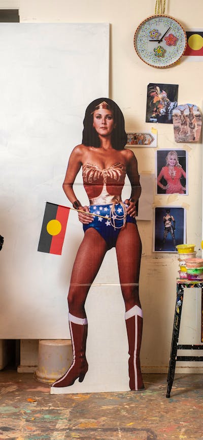 A woman stands in an eclectic art studio, with a vibrant portrait and a Wonder Woman cut-out.