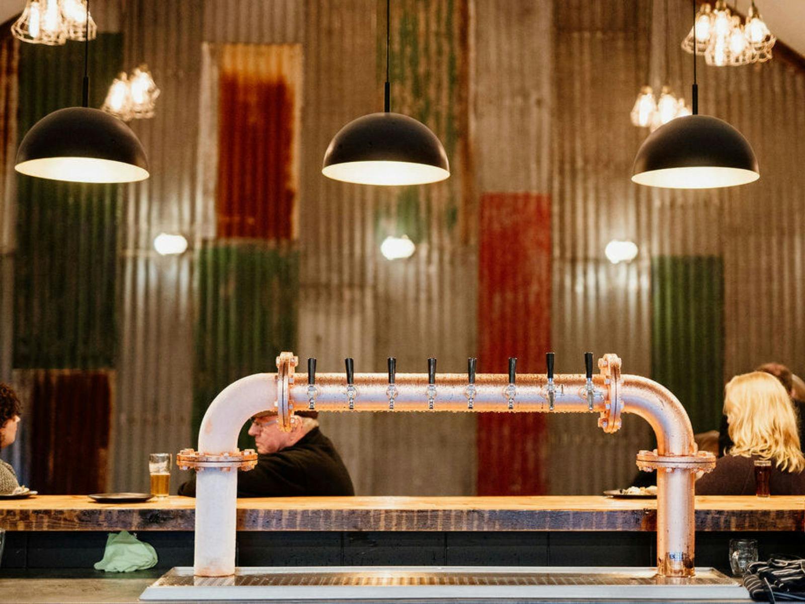 Copper beer taps and patrons seated inside warm industrial-style brewery interior.
