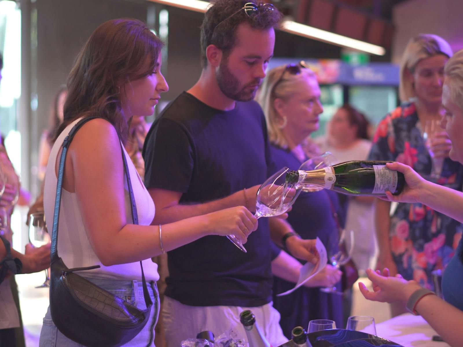 A server pours white wine as a woman and man watch, surrounded by guests in colorful event lighting.