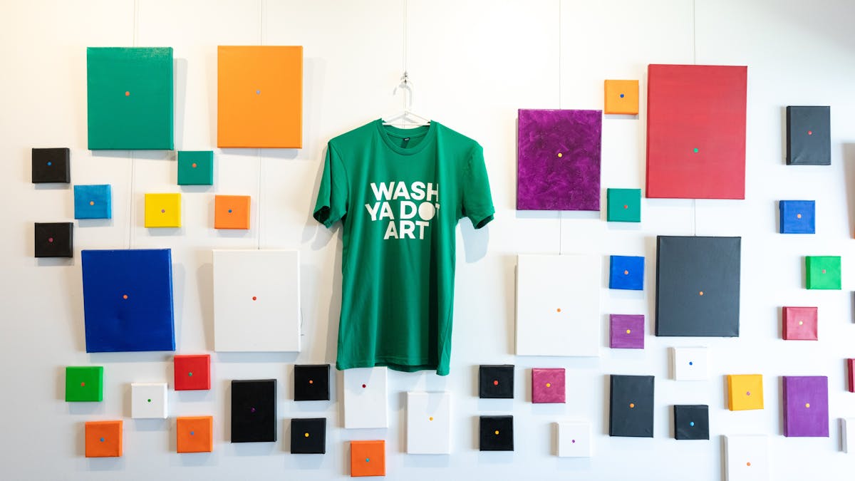 Artwork of a bright green shirt hanging on a wall surrounded by brightly coloured canvases