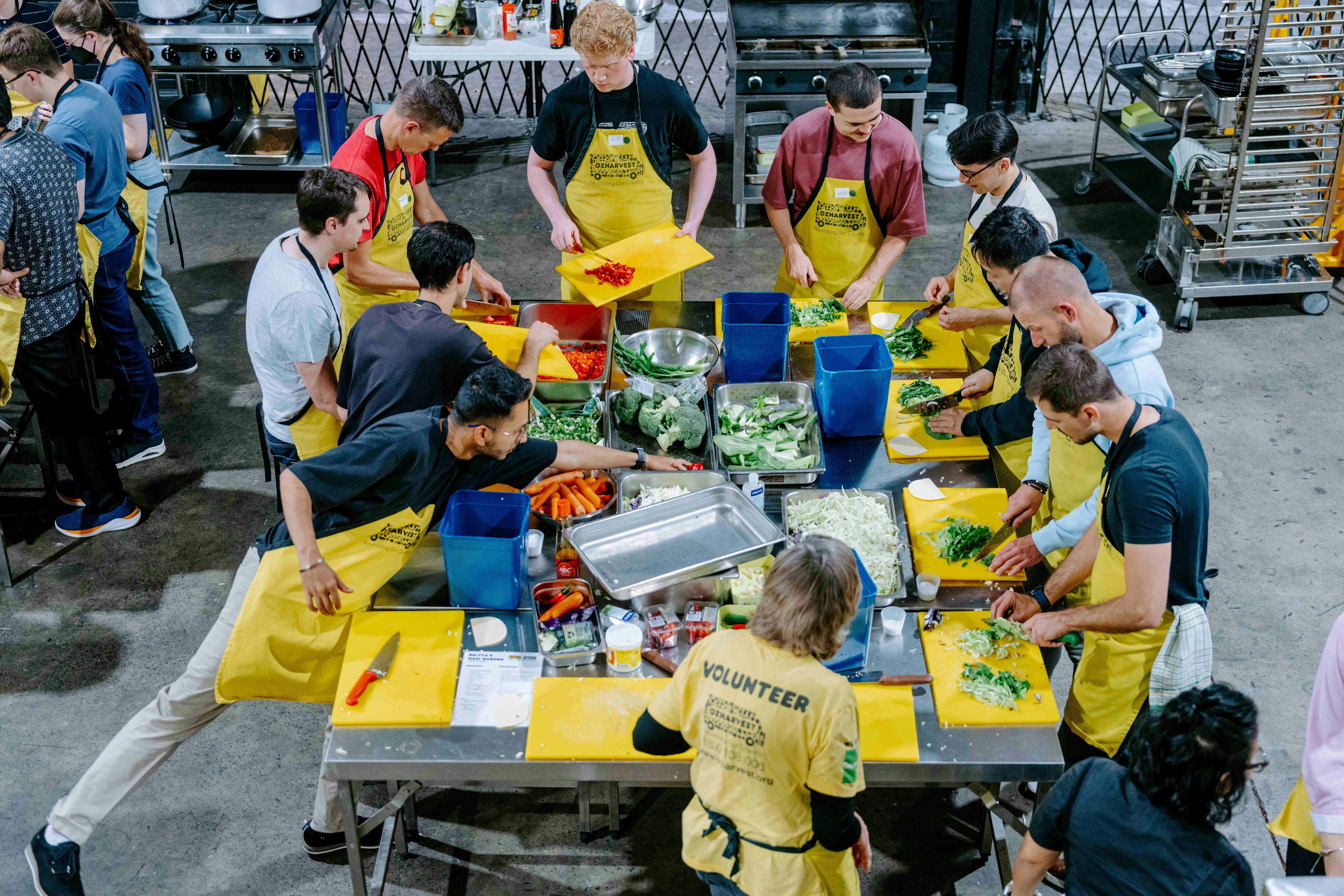 Overhead view of a large group of people in yellow OzHarvest aprons chopping vegetables at a bench