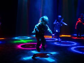 A young child jumping on a circle of light surrounded by other colourful lights and children