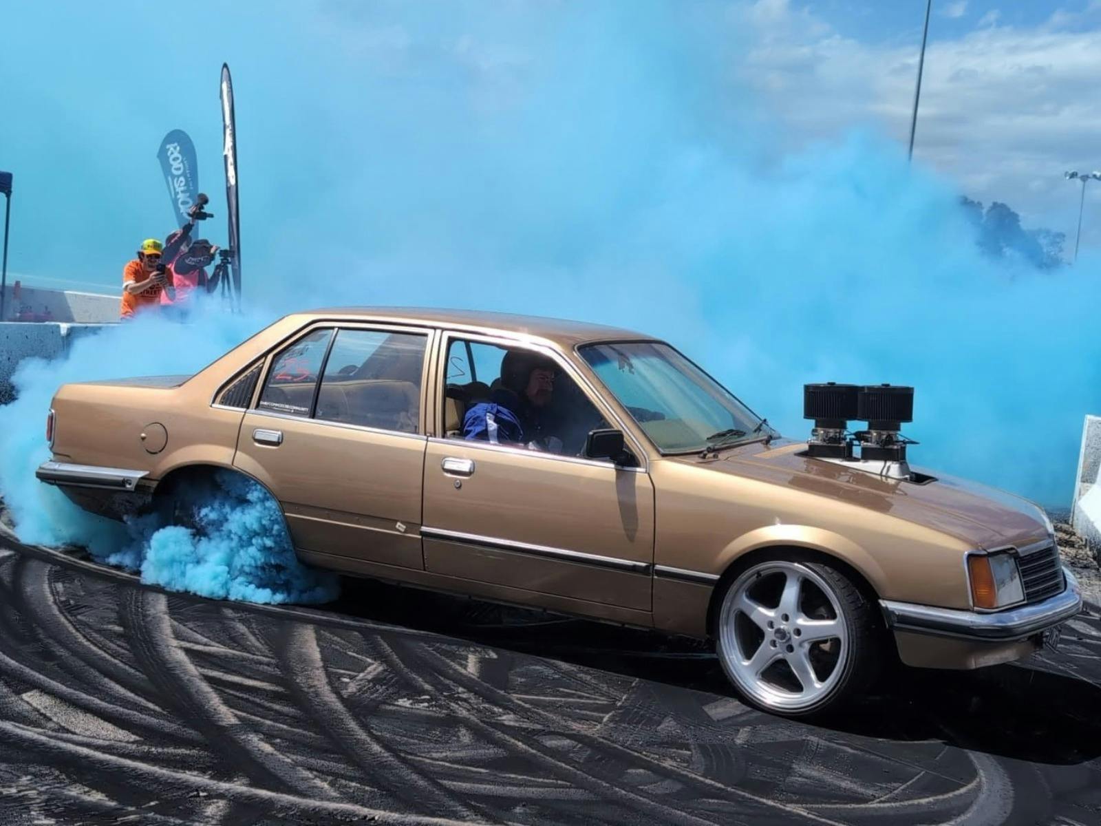 Brown burnout car spinning its rear tyres as blue tyre smoke fills the burnout pad.